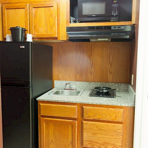 The image shows a small kitchen with wooden cabinets, a microwave, a black refrigerator, a small sink, and a stovetop.