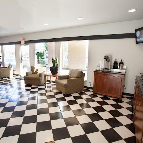 A waiting area with black-and-white checkered flooring, some chairs, a TV, coffee station, and large windows. Walls are white.