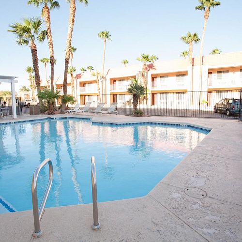 A clear blue swimming pool surrounded by lounge chairs, palm trees, and a building in the background under a bright sky.