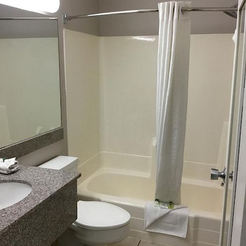 The image shows a clean bathroom with a white tiled floor, a bathtub with a shower curtain, a toilet, a sink, and a large mirror above the countertop.