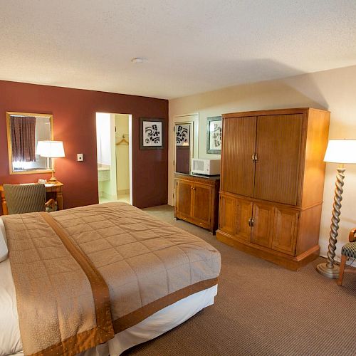 A cozy hotel room with a bed, nightstands, lamps, a chair, two framed pictures, a TV armoire, a mini-fridge, a microwave, and a bathroom entry.