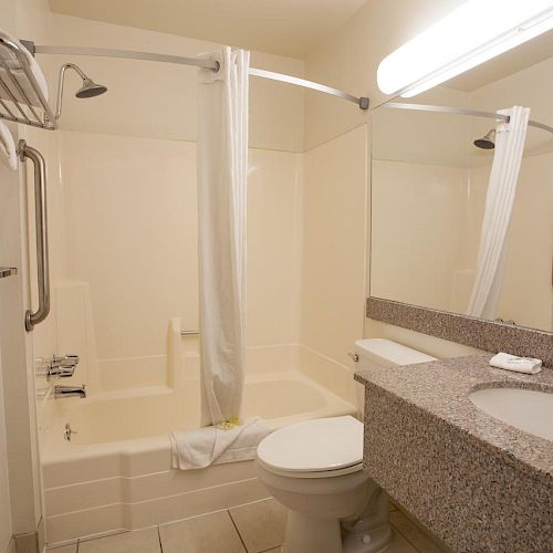 This image shows a clean, white bathroom with a shower-tub combo, a toilet, a large mirror, and a granite countertop with a sink ending the sentence.