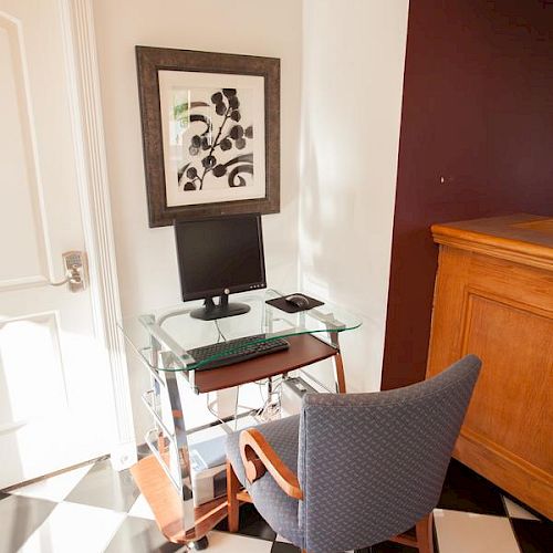 The image shows a small home office with a glass desk, a computer, a framed artwork on the wall, a chair, and checkered black and white floor tiles.