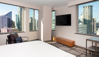 The image shows the corner of a modern apartment or hotel room with city views, featuring large windows, a bed, an armchair, and a wall-mounted TV.