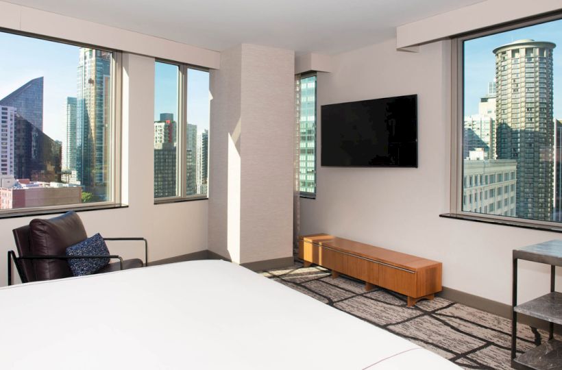 The image shows the corner of a modern apartment or hotel room with city views, featuring large windows, a bed, an armchair, and a wall-mounted TV.