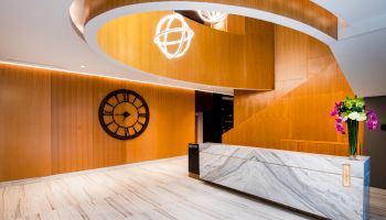 A modern lobby with a marble reception desk, wooden spiral staircase, large wall clock, and a bouquet of flowers on the desk.