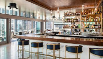 A modern, upscale bar interior with a wooden ceiling, well-stocked shelves, and high-backed chairs; large windows overlook a cityscape outside.