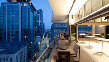 A modern rooftop patio with seating and tables overlooks a cityscape with tall buildings and illuminated streets during the evening.