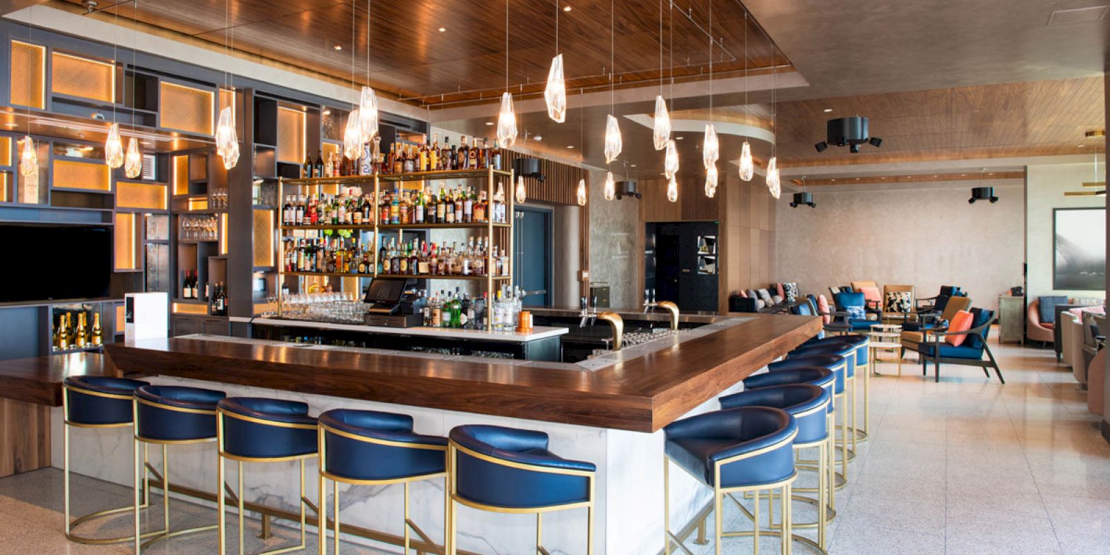 This image shows a modern bar with stylish chairs, a stocked bar, hanging lights, and a cozy seating area in the background.