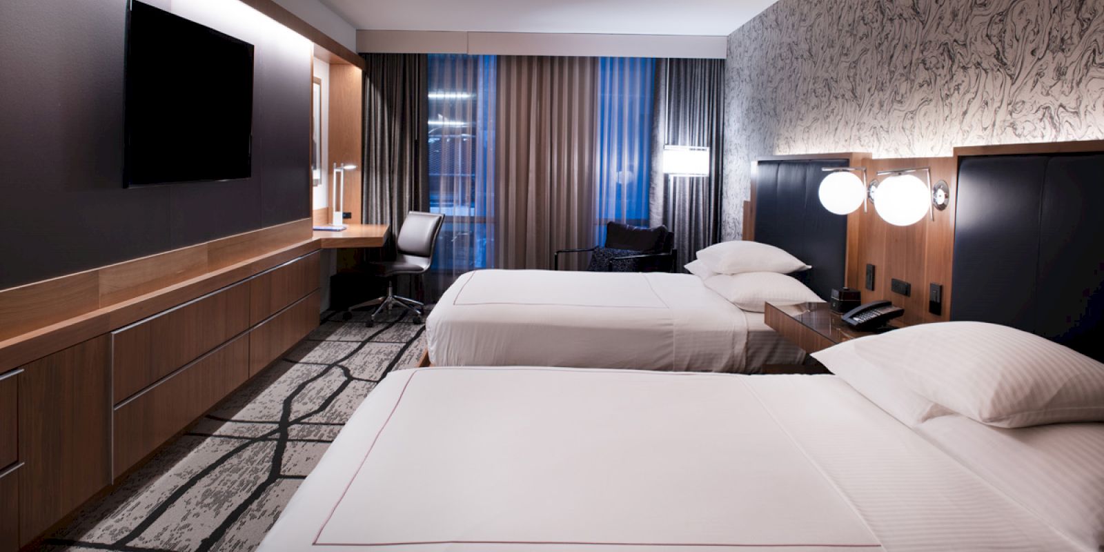 A modern hotel room with two neatly made beds, a large TV, a work desk, and contemporary decor, including textured wallpaper and ambient lighting.