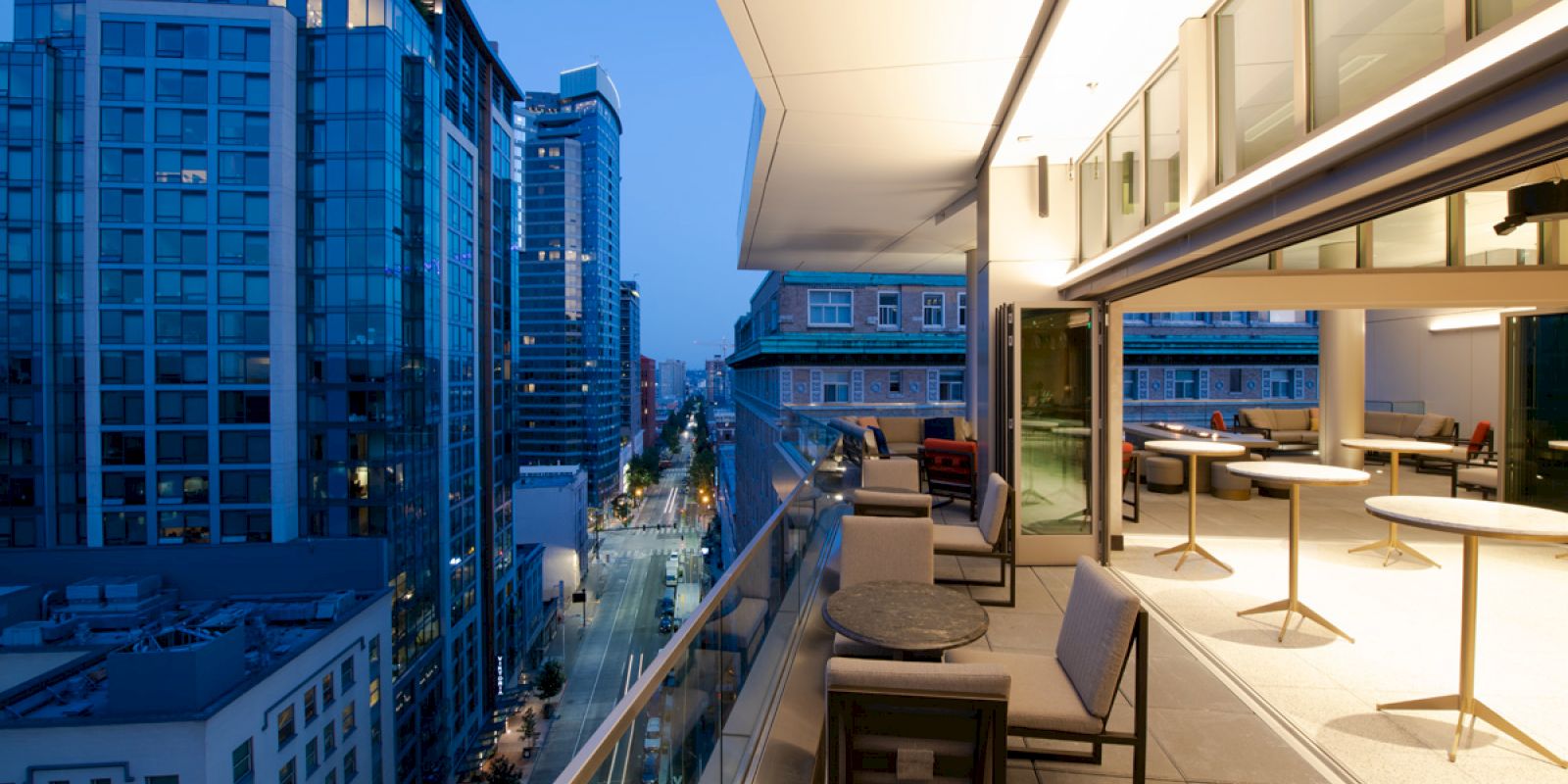 A modern balcony with tables and chairs overlooks a cityscape of tall buildings and streets illuminated in the evening.