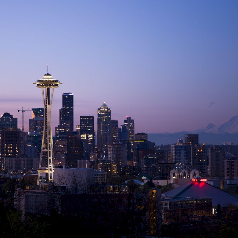 This image shows the skyline of a city at twilight, featuring the Space Needle prominently, with Mount Rainier visible in the background.