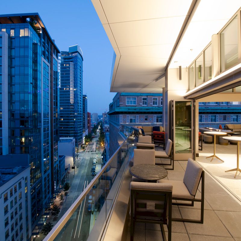 A modern cityscape with tall buildings at dusk, featuring a spacious terrace with seating and tables overlooking the street below.