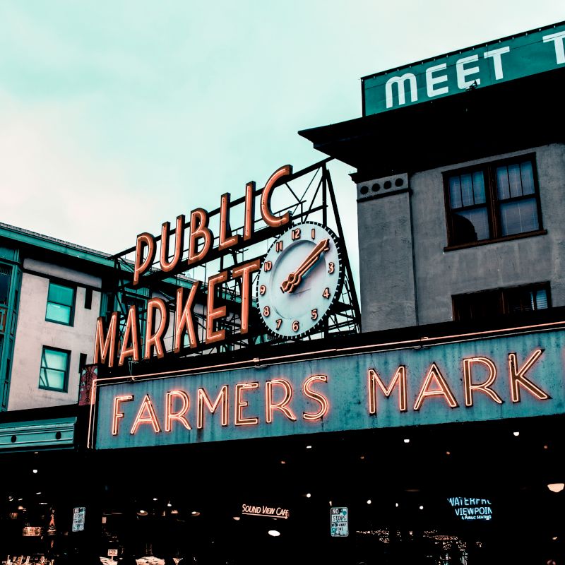 This image shows the entrance to a public market, featuring neon signs that read "Public Market" and "Farmers Market" with a clock between them.