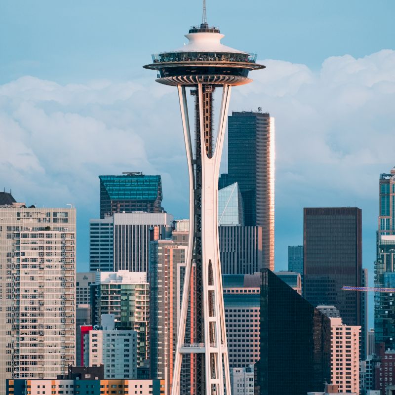 The image shows a cityscape featuring the Space Needle, a prominent tower, surrounded by various high-rise buildings and a blue sky with clouds in the background.