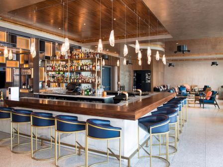 The image shows a modern, stylish bar with high chairs, a well-stocked bar counter, and pendant lighting, located within a spacious lounge area.
