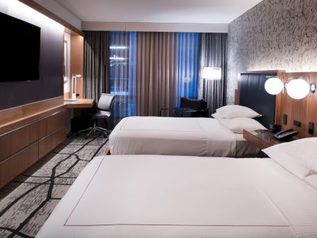 A modern hotel room with two beds, a flat-screen TV, a desk with a chair, and stylish decor featuring patterned wallpaper and contemporary lighting.