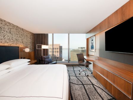 A modern hotel room with a king-sized bed, large TV, desk, chair, and a window view of buildings and water.