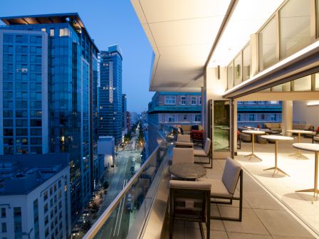 The image shows a modern rooftop terrace with outdoor seating, overlooking a cityscape with tall buildings during evening time.