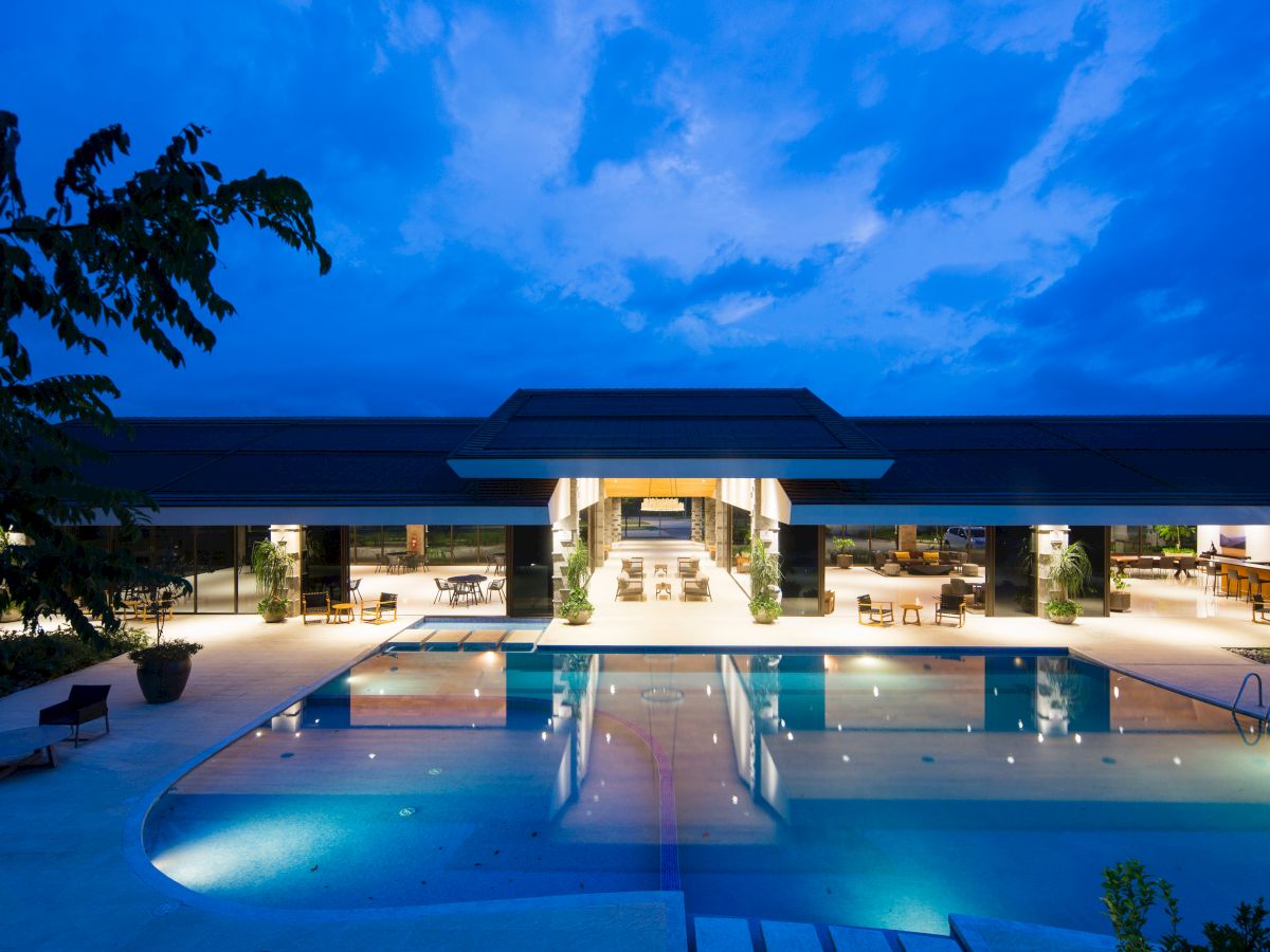 A luxurious poolside view of a well-lit resort at dusk, featuring an open building and clear blue sky.