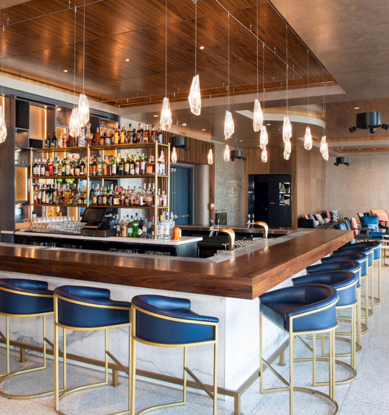 The image shows a modern bar with a wooden counter, blue bar stools, hanging lights, and a well-stocked liquor shelf in a stylish, spacious setting.