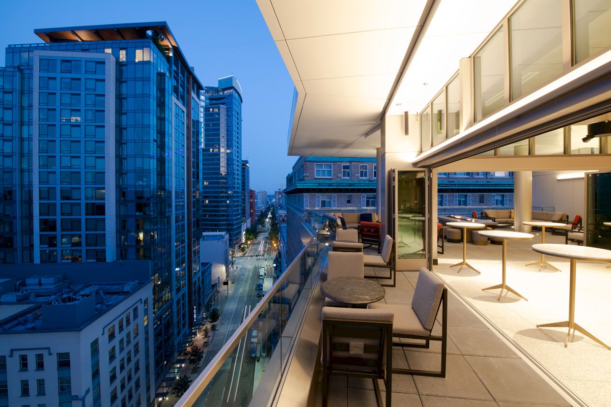 A modern cityscape view from a balcony terrace at dusk, featuring outdoor seating, tall glass buildings, and a slightly illuminated street below.