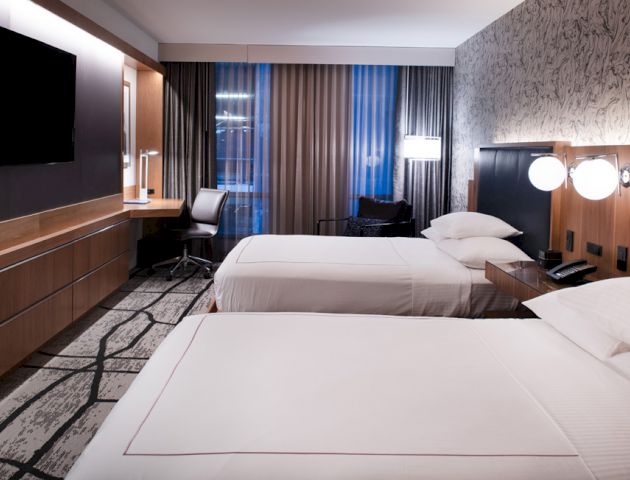 This image shows a modern hotel room with two beds, a flat-screen TV, a desk with a chair, and contemporary decor and lighting.