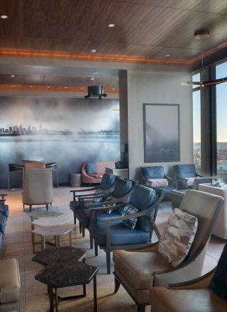 The image shows a modern lounge with comfortable seating, large windows offering a scenic view of the waterfront, and a skyline mural on the wall.
