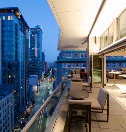 An outdoor terrace with tables and chairs overlooks a city street lined with modern high-rise buildings at dusk.