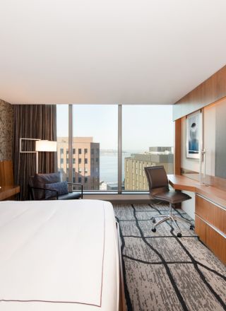 This image shows a modern hotel room with a large bed, desk, chair, TV, and floor-to-ceiling windows offering a view of buildings and water.