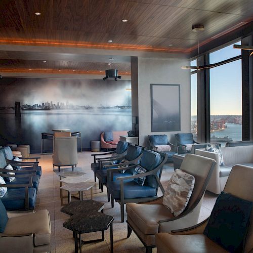 A modern lounge area with comfortable seating, large windows showcasing a scenic view of a waterfront and Ferris wheel, and an artistic skyline mural.