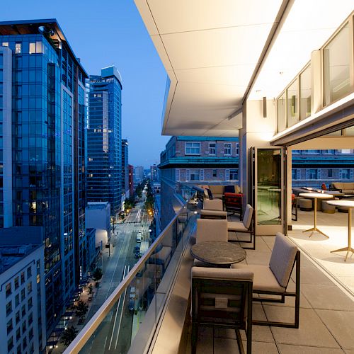 The image shows a modern cityscape at dusk, with tall buildings and an illuminated exterior balcony featuring tables and chairs.