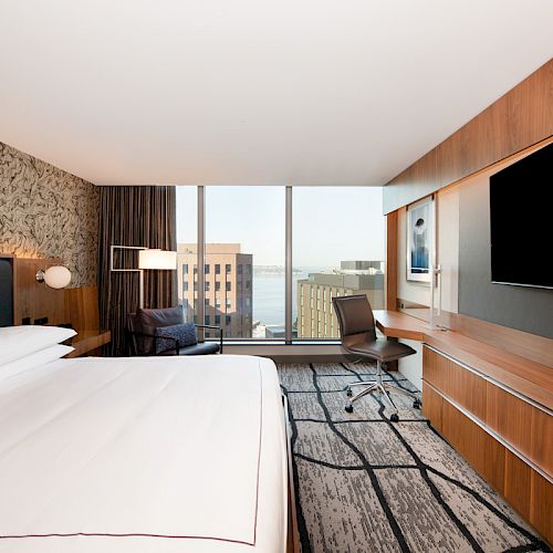 A modern hotel room with a large bed, desk, chair, flat-screen TV, and a city view from large windows.