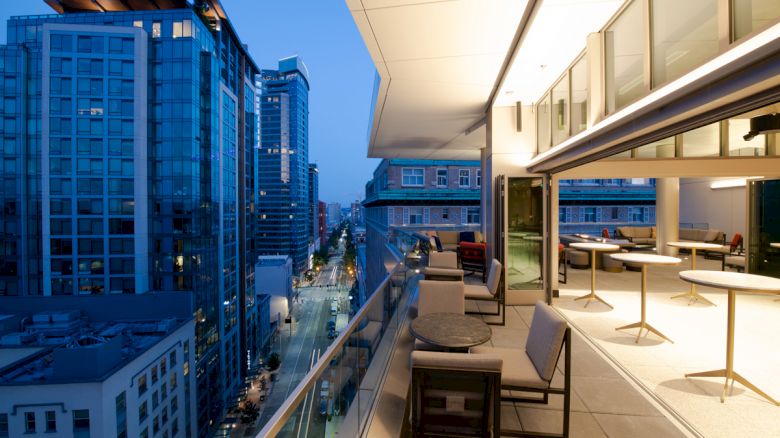 A modern cityscape at dusk with high-rise buildings and a rooftop terrace featuring seating areas, tables, and large windows showing indoor spaces.