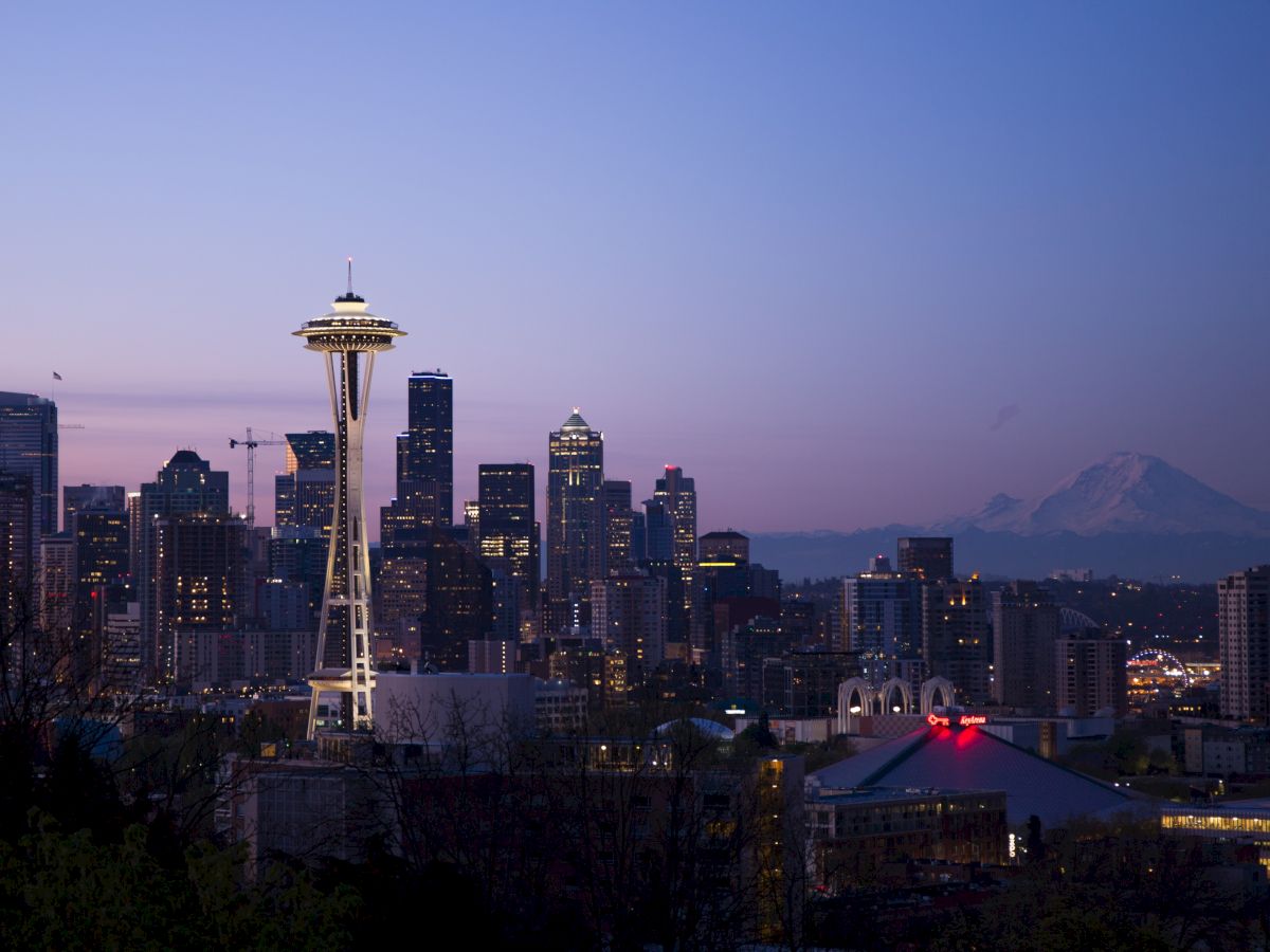 A cityscape at dusk featuring the Space Needle with downtown buildings and Mount Rainier in the background. The sky shows shades of blue and purple.