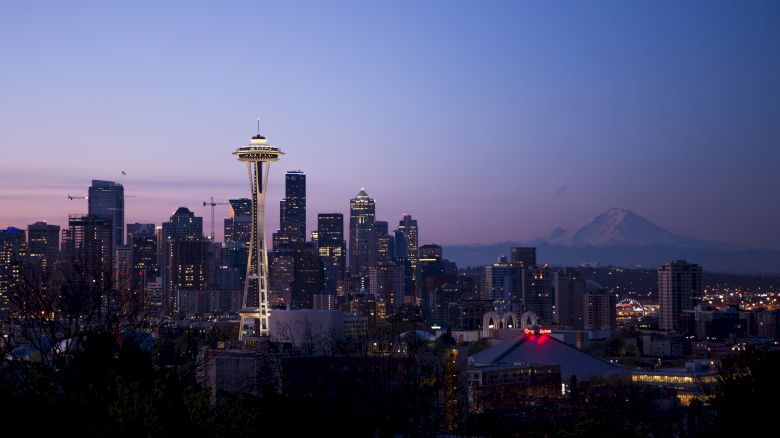 A cityscape at dusk featuring the Space Needle with downtown buildings and Mount Rainier in the background. The sky shows shades of blue and purple.