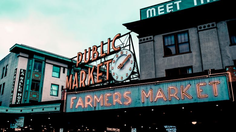 An outdoor scene shows signs for a Public Market, Farmers Market, and a retro clock. Buildings and a hotel are in the background.
