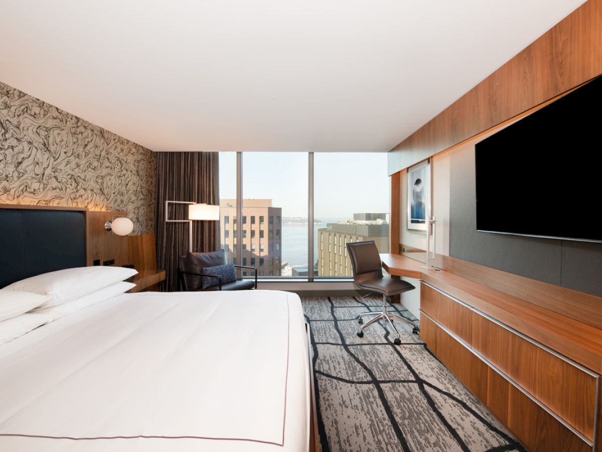 A modern hotel room with a large bed, wall-mounted TV, desk, chair, and a window offering a city view. The décor includes wood tones and a patterned wall.