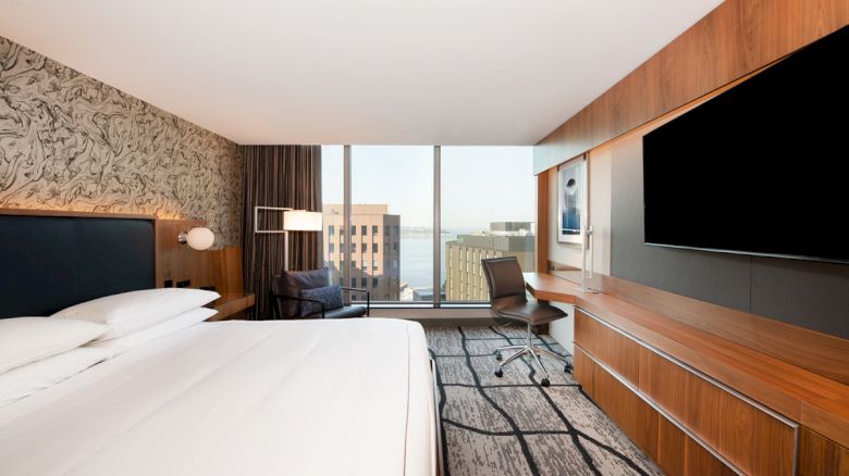A modern hotel room with a large bed, wall-mounted TV, desk, chair, and a window offering a city view. The décor includes wood tones and a patterned wall.