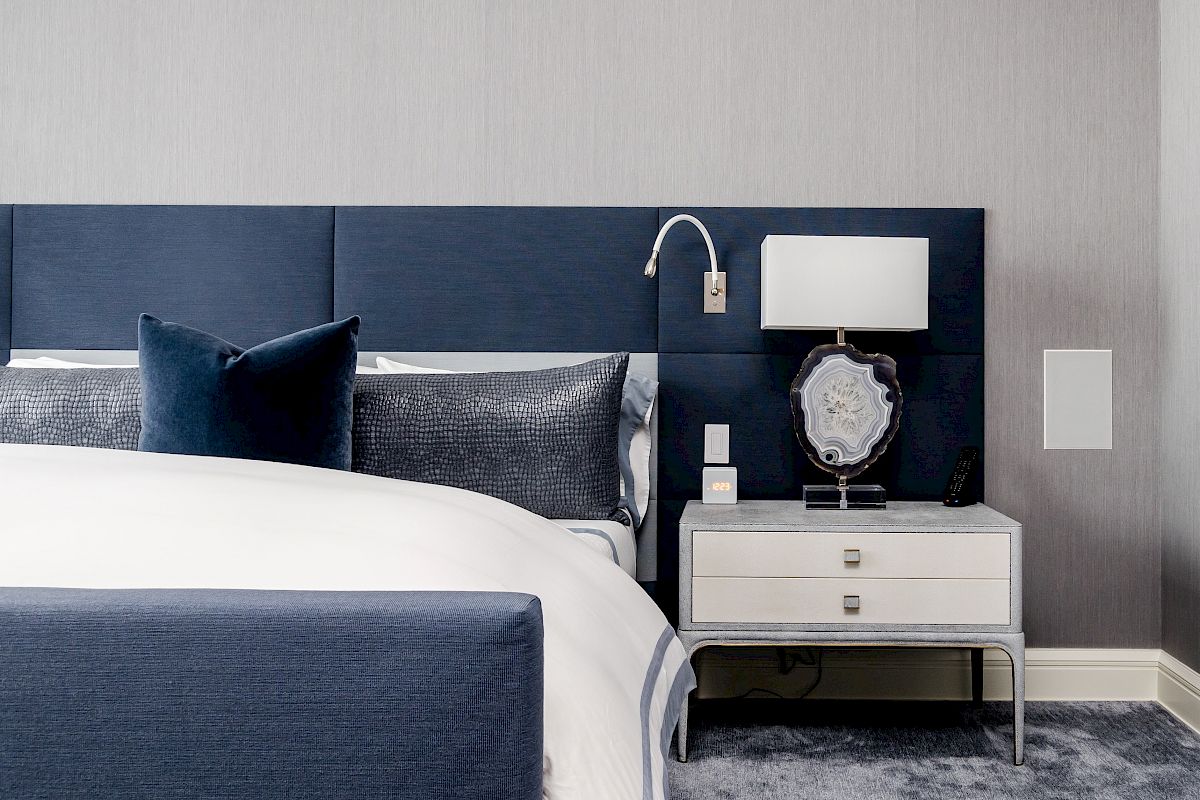 A modern bedroom with a blue headboard, white and blue bedding, a nightstand with a lamp, and a decorative object on a gray carpet.