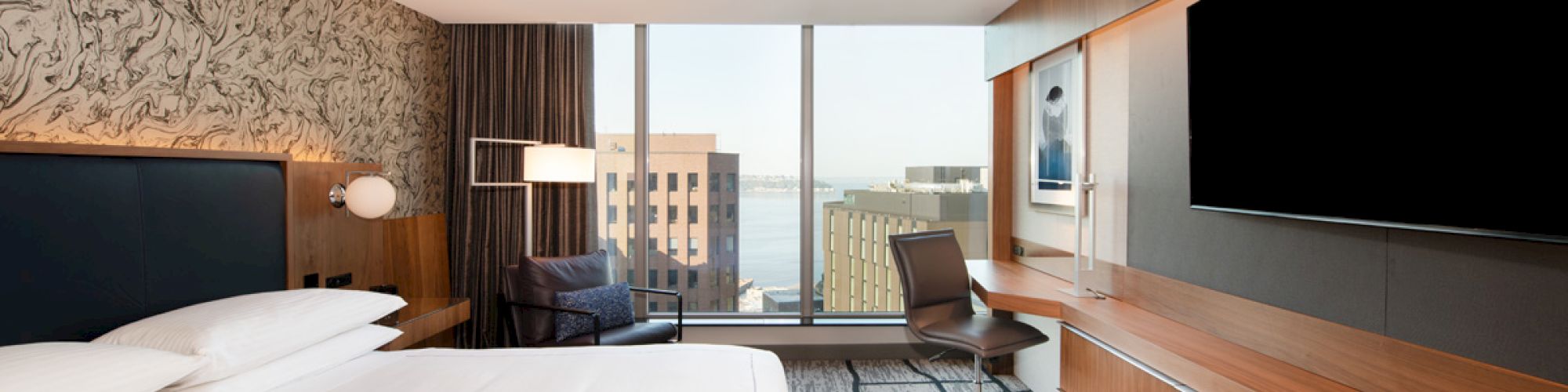 A modern hotel room with a large bed, a wall-mounted TV, a desk with a chair, and a window with a city view is shown.